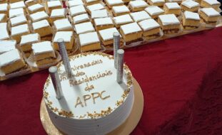 Fotografia de um bolo de aniversário, redondo, pousado numa mesa com uma toalha de tom roxo. Junto do pequeno bolo também várias pequenas fatias, em formato quadrado, de um bolo previamente cortado.