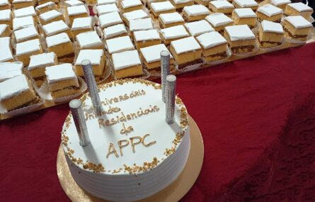 Fotografia de um bolo de aniversário, redondo, pousado numa mesa com uma toalha de tom roxo. Junto do pequeno bolo também várias pequenas fatias, em formato quadrado, de um bolo previamente cortado.