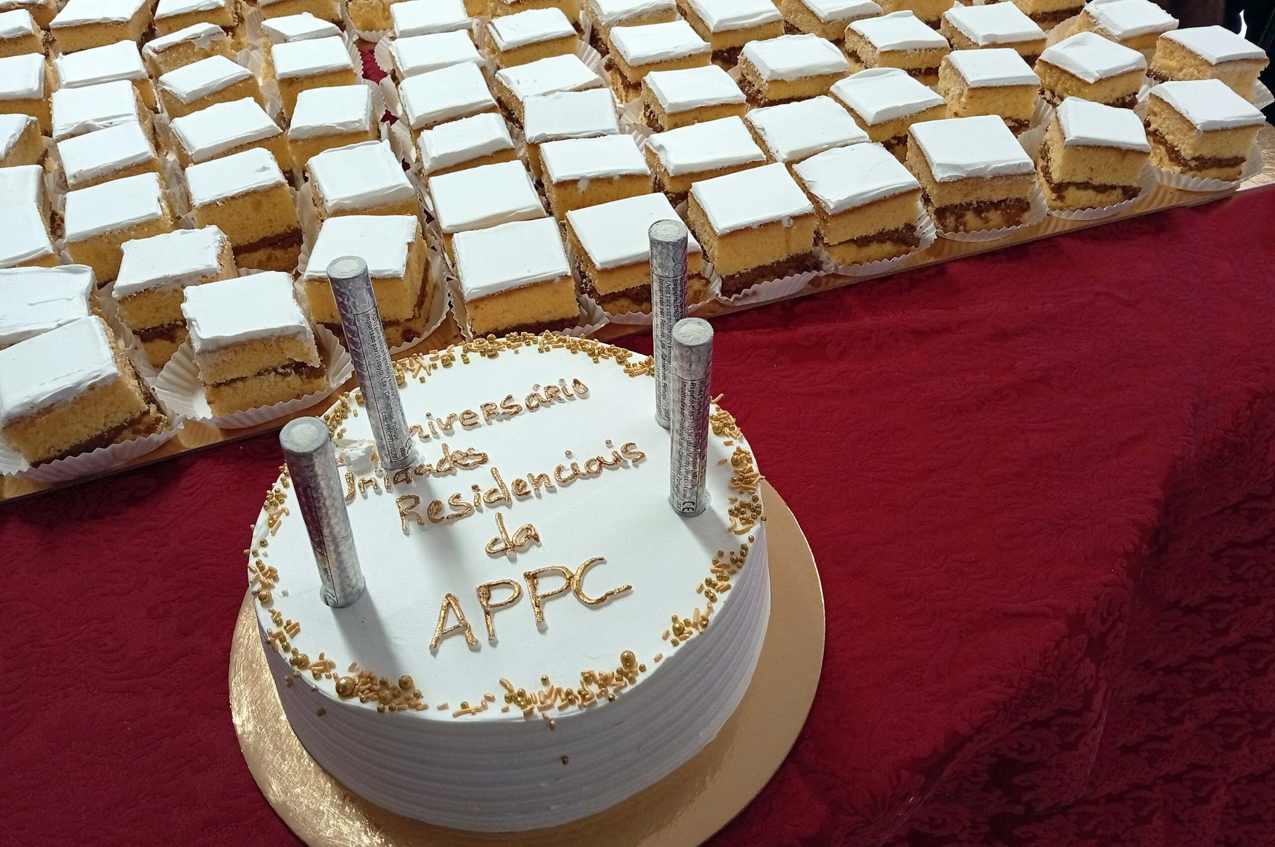 Fotografia de um bolo de aniversário, redondo, pousado numa mesa com uma toalha de tom roxo. Junto do pequeno bolo também várias pequenas fatias, em formato quadrado, de um bolo previamente cortado.