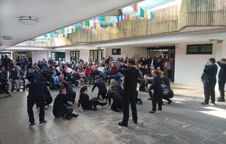 Espaço exterior com várias pessoas (sentadas e em pé) a assistirem a uma atuação de um grupo de dança.