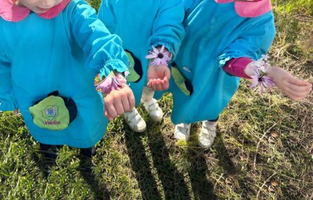Três crianças em espaço relvado. A fotografia foi realizada numa perspetiva superior e as três crianças têm os braços esquerdos estendidos e mostram, no pulso, pequenas pulseiras feitas com diversas flores.