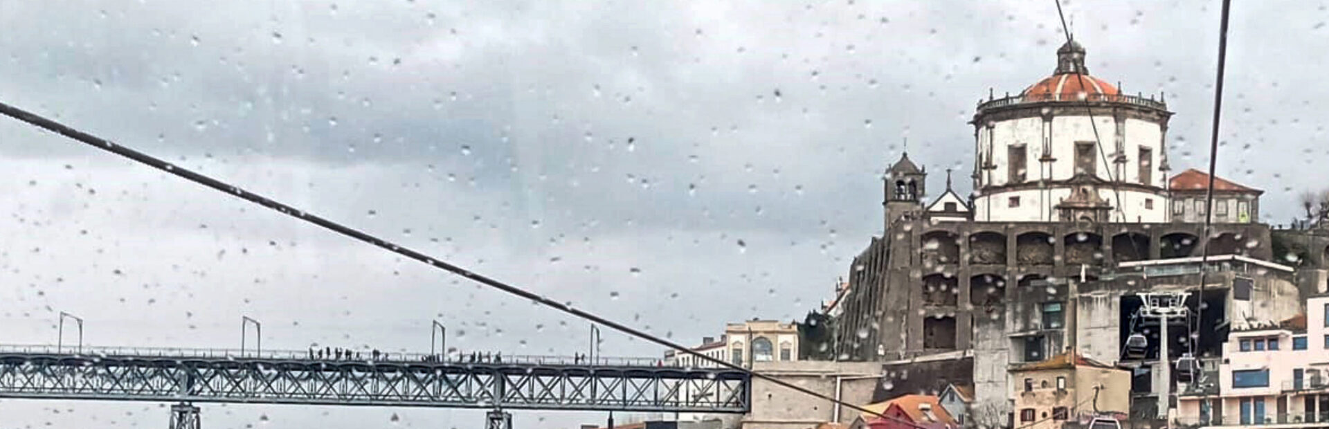 Imagem de parte da zona ribeirinha de Vila Nova de Gaia. São visíveis inúmeros edifícios (essencialmente antigos) e, ainda que parcialmente, duas pontes. Na imagem surgem ainda cinco cabines de teleférico e os respetivos cabos de transporte das mesmas.