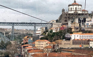 Imagem de parte da zona ribeirinha de Vila Nova de Gaia. São visíveis inúmeros edifícios (essencialmente antigos) e, ainda que parcialmente, duas pontes. Na imagem surgem ainda cinco cabines de teleférico e os respetivos cabos de transporte das mesmas.