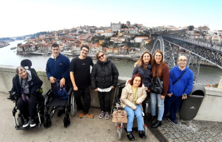 Imagem de oito pessoas, lado a lado. Atrás e ao fundo surge uma ponte (em metal), imensos edifícios e é visível um pouco de rio. No rio, atracados junto à margem, estão vários barcos.