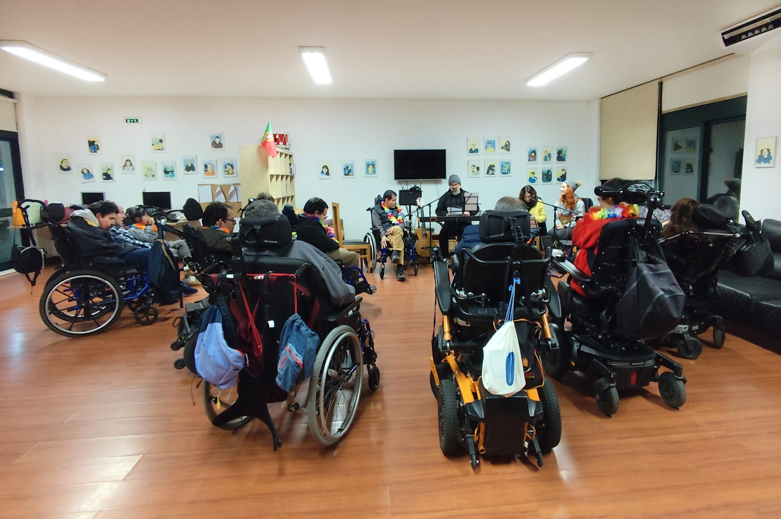 Fotografia de espaço interior com cerca de uma dezena de pessoas. Em primeiro plano, de costas, estão várias pessoas (em cadeira de rodas) em formato de semicírculo. Ao fundo, de frente, surgem três pessoas junto de microfones e outros equipamentos de som. A parede do fundo está repleta de pequenas ilustrações de várias pessoas.