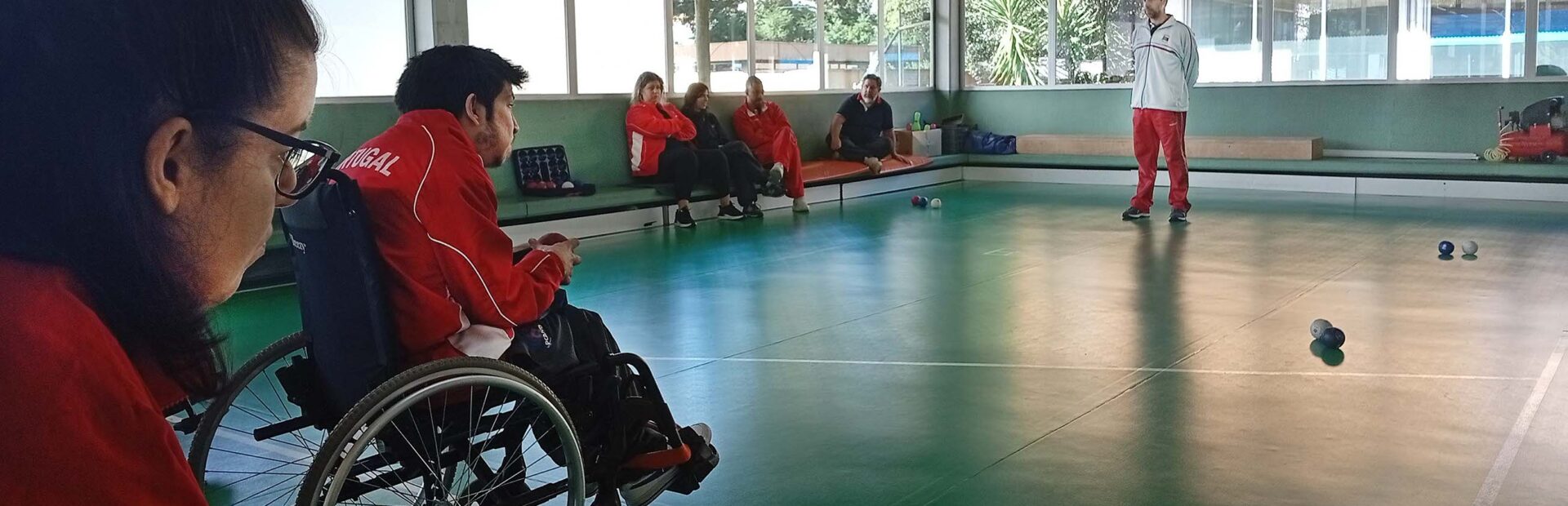 Fotografia de ginásio interior. Dois atletas de boccia em primeiro plano. Ao fundo da imagem estão quatro pessoas sentadas e uma pessoa em pé. Várias bolas de boccia espalhadas pelo chão.