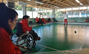 Fotografia de ginásio interior. Dois atletas de boccia em primeiro plano. Ao fundo da imagem estão quatro pessoas sentadas e uma pessoa em pé. Várias bolas de boccia espalhadas pelo chão.