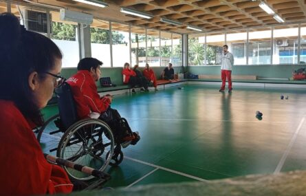 Fotografia de ginásio interior. Dois atletas de boccia em primeiro plano. Ao fundo da imagem estão quatro pessoas sentadas e uma pessoa em pé. Várias bolas de boccia espalhadas pelo chão.