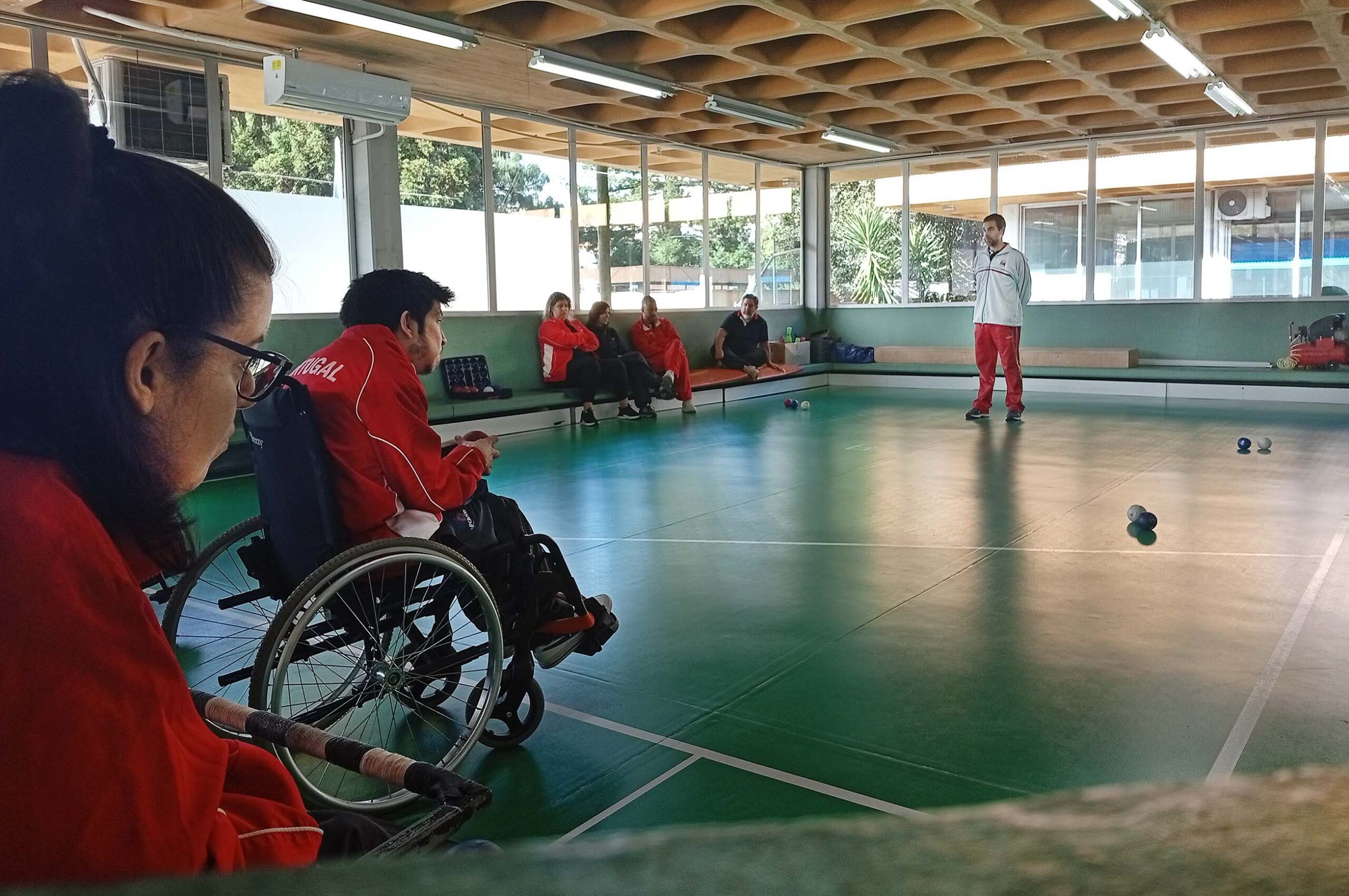 Fotografia de ginásio interior. Dois atletas de boccia em primeiro plano. Ao fundo da imagem estão quatro pessoas sentadas e uma pessoa em pé. Várias bolas de boccia espalhadas pelo chão.