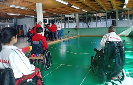 Fotografia de ginásio interior. Três atletas de boccia, de costas, em primeiro plano. Ao fundo da imagem estão várias pessoas sentadas. São visíveis cinco bolas de boccia espalhadas pelo chão.