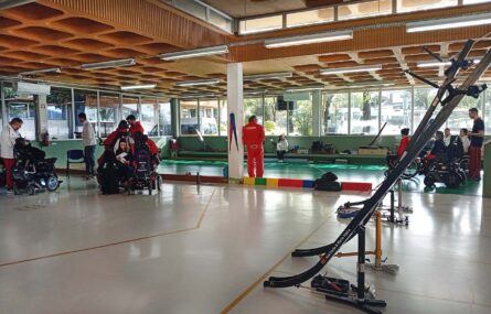 Fotografia ampla de ginásio interior. Várias pessoas preparam-se para a prática de boccia e, também, algumas assistem. São visíveis várias calhas de boccia em primeiro plano.