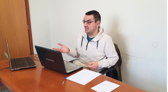 Pessoa sorridente, está sentada em frente a computador. Sobre a mesa estão também dispostos alguns papeis de forma organizada.
