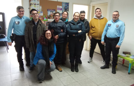 Grupo de nove pessoas em espaço interior de edifício. Três das pessoas do grupo usam indumentária de força de segurança (Polícia de Segurança Pública). Todos estão sorridentes. Na parede, atrás das pessoas, está um quadro com vários cartazes e folhas com informações diversas.