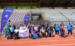 Perto de três dezenas de pessoas fotografadas na pista de atletismo de um espaço desportivo exterior. Junto do grupo estão dois estandartes azuis com a designação “Maia Desporto”. Atrás aparecem as bancadas.