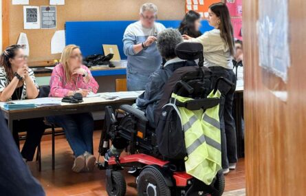 Fotografia de duas pessoas junto de uma mesa de voto, em sala interior. Do outro lado da mesa estão cinco pessoas – sendo que os rostos dessas pessoas foram digitalmente desfocados. Inúmeros papéis afixados em quadro na parede e diversos documentos pousados em cima da mesa de votação.