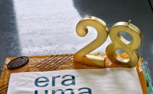 Fotografia de um bolo. O bolo tem duas velas em forma de algarismos com os números dois e oito em tons dourados. Junto dos algarismos está uma imagem com o logótipo do “Era uma vez... Teatro”. O bolo tem ainda (no canto superior direito) pousado um pedaço de chocolate em formato oval.