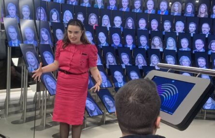 Pessoa de vestido rosa de pé em frente a uma audiência. O fundo consiste em várias telas a exibir rostos. Outra pessoa está sentada e voltada para a pessoa de vestido vermelho, com um tablet ou dispositivo de ecrã visível à sua frente.