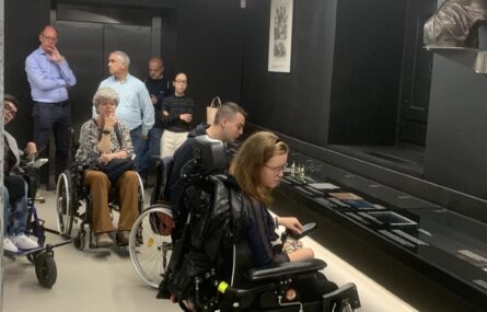 Grupo de pessoas, algumas em cadeiras de rodas, reunidas num museu ou galeria. Estão de frente para uma vitrine e um busto num pedestal. A sala tem paredes escuras e é iluminada por luzes no teto.