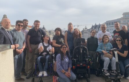 Grupo de pessoas, incluindo utilizadores de cadeira de rodas, posa para uma fotografia no exterior com vista para edifícios históricos. O ambiente é descontraído e alegre.