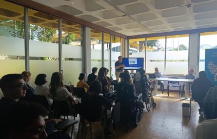 Sala de conferências com várias pessoas sentada. Há um orador em pé ao fundo, próximo a uma tela que exibe uma apresentação. A sala tem grandes janelas com vidro fosco e luz natural a entrar..