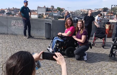 Um grupo de pessoas ao ar livre numa calçada. Uma pessoa em primeiro plano está a tirar uma foto com um smartphone. Ao fundo, há três pessoas a posar para a foto, uma das quais usa uma cadeira de rodas. O cenário parece ser uma área urbana com edifícios ao longe.