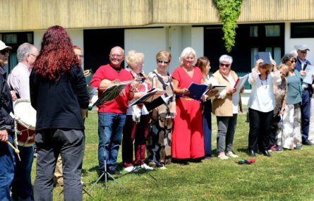Grupo de 14 pessoas em relvado exterior. Algumas pessoas têm instrumentos musicais nas mãos e outras seguram pequenos cadernos com folhas.