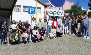 Grupo de 29 pessoas em espaço exterior junto de um edifício. Na parte frontal desse edifício está uma placa azul, redonda, e uma outra com as letras APCV.