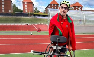 Fotografia de uma pessoa em pista de atletismo de estádio. São ainda visíveis as várias pistas e, ao fundo da imagem, alguns edifícios. A pessoa fotografada tem junto de si uma tricicleta (parcialmente visível).
