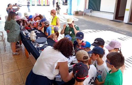 Cerca de duas dezenas de crianças e cinco pessoas adultas junto de duas mesas com material diverso espalhado. Ao fundo da imagem surgem mais alguns adultos. Os rostos visíveis das crianças foram digitalmente desfocados.