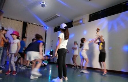Mais de uma dezena de crianças a dançar em sala interior pouco iluminada e com vários focos luminosos. Os rostos visíveis das crianças foram digitalmente desfocados.