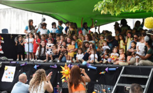 Várias dezenas de crianças e alguns adultos em cima de um palco (com um toldo verde). À frente do palco estão três adultos, de joelhos, a filmar ou fotografar usando telemóveis. Os rostos visíveis das crianças foram digitalmente desfocados.
