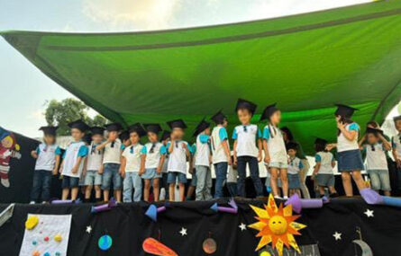 Cerca de duas dezenas de crianças em cima de um palco (com um toldo verde). Todas as crianças usam uma cartola negra. A zona frontal do palco está repleta de adereços alusivos ao Sistema Solar. Os rostos visíveis das crianças foram digitalmente desfocados.
