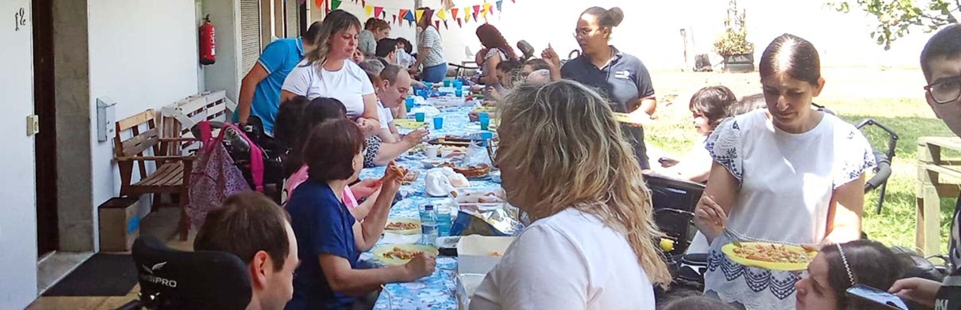 Várias pessoas em convívio junto de uma mesa comprida colocada em espaço exterior. A mesa está repleta de alimentos e bebidas.