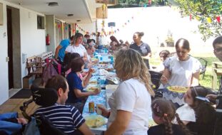 Várias pessoas em convívio junto de uma mesa comprida colocada em espaço exterior. A mesa está repleta de alimentos e bebidas.