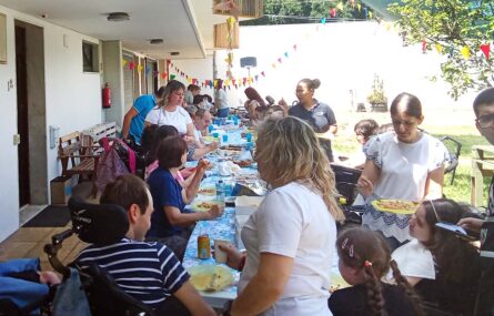 Várias pessoas em convívio junto de uma mesa comprida colocada em espaço exterior. A mesa está repleta de alimentos e bebidas.