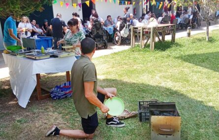 Várias pessoas em convívio junto de uma mesa comprida colocada em espaço exterior. Em primeiro plano está uma pessoa ajoelhada num espaço relvado (junto de um pequeno fogareiro).