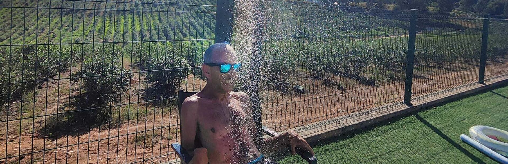 Uma pessoa sentada numa cadeira, num jardim, está debaixo de um chuveiro do qual sai imensa água. Ao fundo da imagem terrenos cultivados e várias árvores.