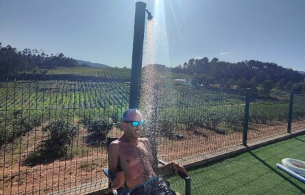 Uma pessoa sentada numa cadeira, num jardim, está debaixo de um chuveiro do qual sai imensa água. Ao fundo da imagem terrenos cultivados e várias árvores.