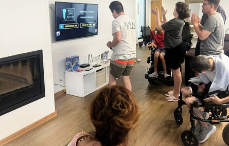 Grupo de pessoas em sala interior. Na parede está um ecrã com imagens de um programa digital de karaoke.