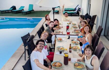 Grupo de 11 pessoas sentadas junto de duas mesas em espaço exterior ao lado de uma piscina. A mesa tem inúmeros alimentos e bebidas.