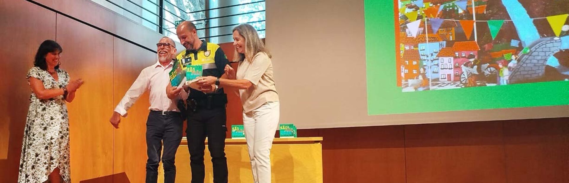 Quatro pessoas em cima de um palco. Atrás dessas pessoas uma projeção multimédia em tela gigante. As quatro pessoas estão claramente sorridentes e uma delas segura nas mãos o que aparenta ser um prémio ou troféu.