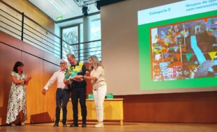 Quatro pessoas em cima de um palco. Atrás dessas pessoas uma projeção multimédia em tela gigante. As quatro pessoas estão claramente sorridentes e uma delas segura nas mãos o que aparenta ser um prémio ou troféu.