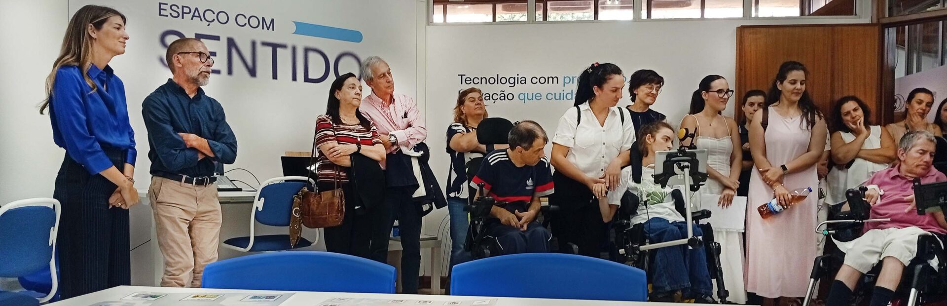 Mais de uma dezena de pessoas em sala apetrechada com diverso equipamento informático. Em primeiro plano uma mesa com diversas tabelas de comunicação pousadas. Numa das paredes, com letras grandes, surge a frase “Espaço com Sentido”.