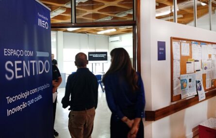 Duas pessoas, de costas e em contraluz, entram numa sala. Em primeiro plano um cartaz com indicação “Fundação MEO” e “Espaço com Sentido. Tecnologia com propósito. Inovação que cuida.