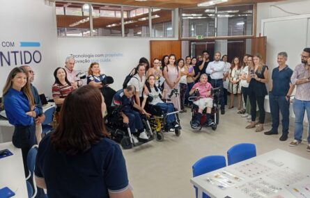 Mais de duas dezenas de pessoas em sala apetrechada com diverso equipamento informático. Em primeiro plano uma mesa com diversas tabelas de comunicação pousadas. Numa das paredes está a inscrição “Tecnologia com propósito. Inovação que cuida.”