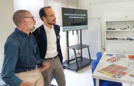 Duas pessoas conversam em espaço interior. Vê-se um ecrã com uma projeção, prateleiras com inúmero material informático e tecnológico e, ainda, uma mesa com tabelas de comunicação pousadas.