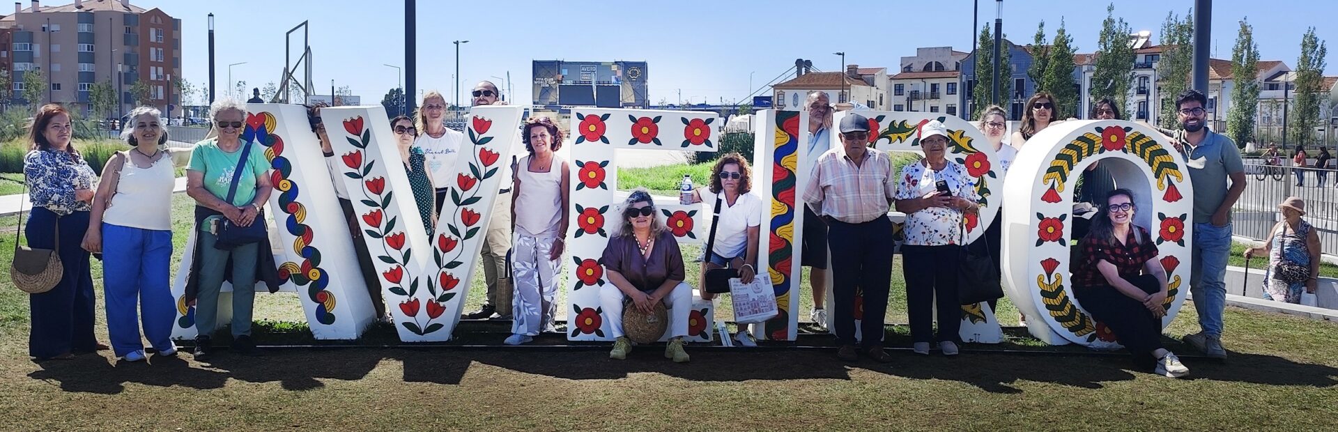 Grupo de 17 pessoas em espaço exterior junto de um placard que refere “Cruzeiros na Ria de Aveiro”. Céu azul e edifícios ao fundo da imagem.