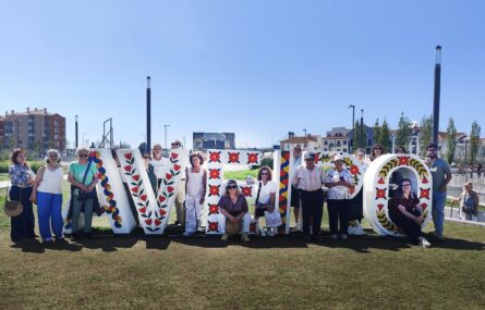 Grupo de 17 pessoas em espaço exterior junto de um placard que refere “Cruzeiros na Ria de Aveiro”. Céu azul e edifícios ao fundo da imagem.