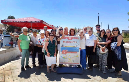Grupo de 17 pessoas em espaço exterior junto de um placard que refere “Cruzeiros na Ria de Aveiro”. Céu azul e edifícios ao fundo da imagem.