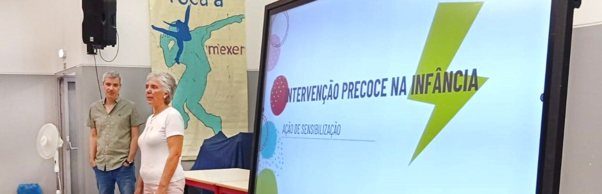 Uma pessoa aparenta estar a falar. Ao lado está um ecrã com uma projeção na qual se lê “Como é que as crianças se desenvolvem?”. A restante informação, no ecrã, é ilegível.
